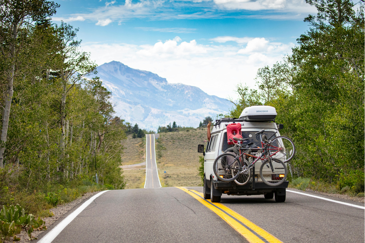 A gray van with bikes strapped to the back driving away from the camera down a scenic highway with a mountain in the distance
